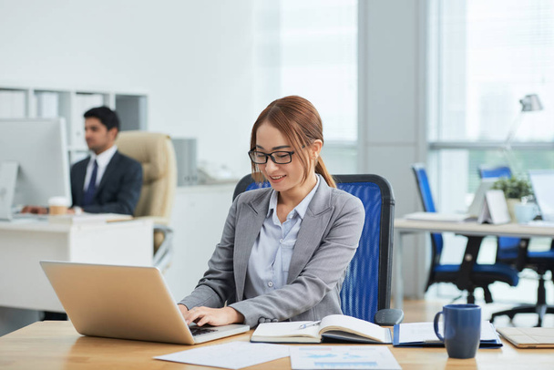 Lächelnde junge Sekretärin mit Brille sitzt am Tisch und tippt im Büro am Laptop, während im Hintergrund ein Geschäftsmann arbeitet - Foto, Bild