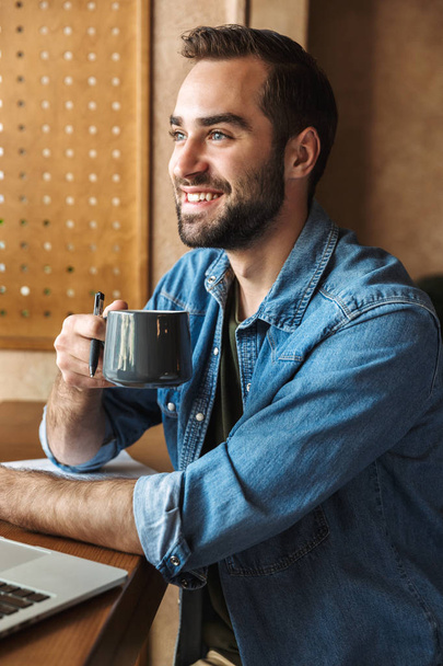 Photo of happy bearded man shirt drinking coffee while working in cafe indoors - Фото, зображення
