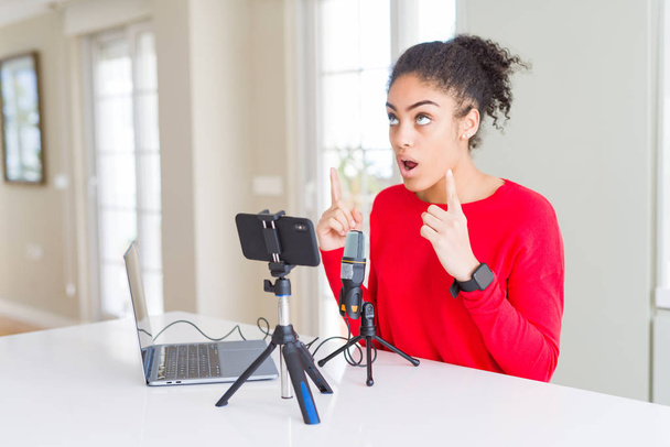 Jeune femme afro-américaine faisant un appel vidéo à l'aide d'une caméra smartphone et d'un microphone étonné et surpris de regarder vers le haut et pointant avec les doigts et les bras levés
. - Photo, image