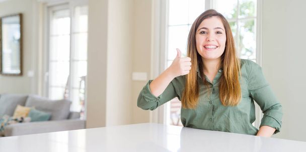 Beautiful young woman at home doing happy thumbs up gesture with hand. Approving expression looking at the camera showing success. - Photo, Image
