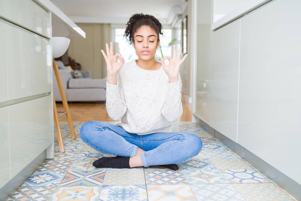 Gyönyörű fiatal afro-amerikai nő afro hajú ül a padlón pihenni, és mosolyogva csukott szemmel csinál meditációs gesztus ujjal. Jóga-koncepció. - Fotó, kép