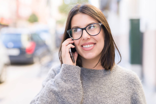 junge schöne Frau mit Brille lächelt selbstbewusst am Telefon und geht auf der Straße in der Stadt - Foto, Bild