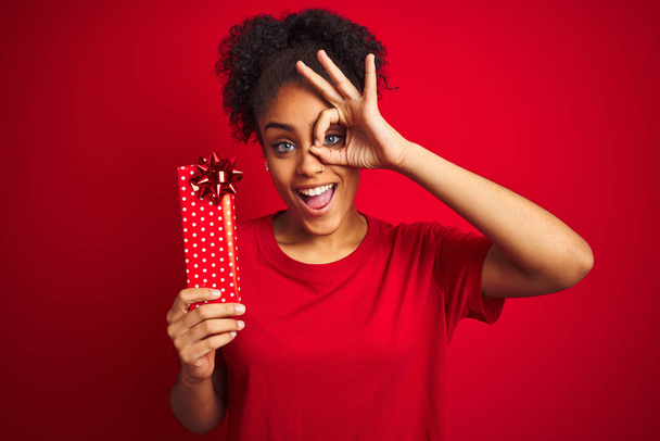 孤立した赤い背景の上にバレンタインギフトを持つ若いアフリカ系アメリカ人女性は、指を通して目に手を当てて笑顔で笑顔を浮かべて - 写真・画像