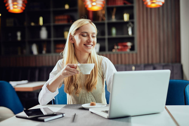 Encantadora mujer de negocios rubia sonriente vestida elegante informal sentada en la cafetería, bebiendo café y utilizando el ordenador portátil
. - Foto, imagen