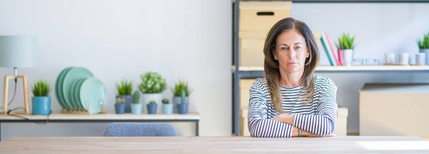Wide angle photo of middle age senior woman sitting at the table at home skeptic and nervous, disapproving expression on face with crossed arms. Negative person. - Photo, Image