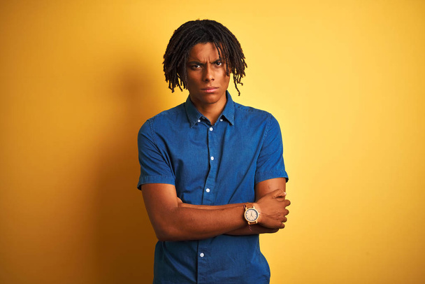 Afro man with dreadlocks wearing casual denim shirt standing over isolated yellow background skeptic and nervous, disapproving expression on face with crossed arms. Negative person. - Photo, Image