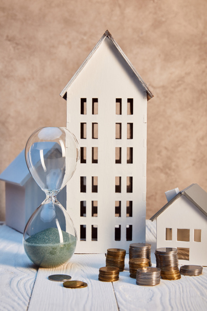 houses models, coins and hourglass on white wooden table near textured wall, real estate concept - 写真・画像