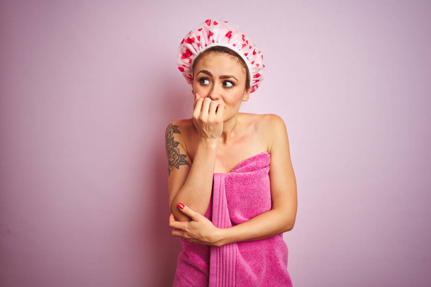 ピンクの孤立した背景の上にシャワーの後にタオルとバス帽子を身に着けている若い美しい女性は、口を噛む爪に手でストレスと緊張を見ています。不安の問題. - 写真・画像