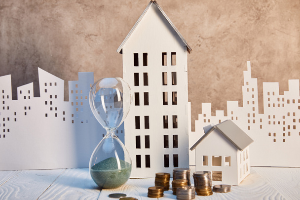 houses models and hourglass on white wooden table with coins, real estate concept - Photo, Image