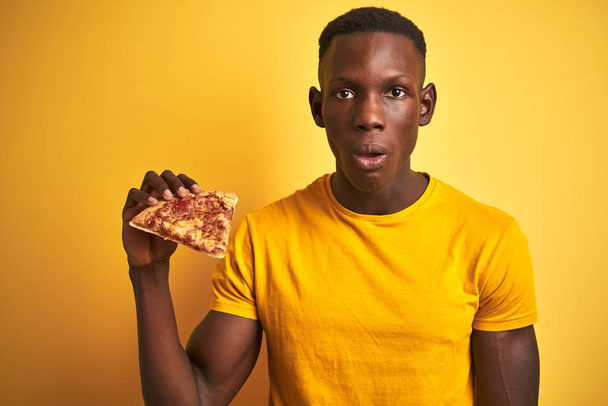 Afro-amerikai ember eszik szelet pizza áll át elszigetelt sárga háttér félek sokk egy meglepetés arc, fél és izgatott a félelem kifejezése - Fotó, kép