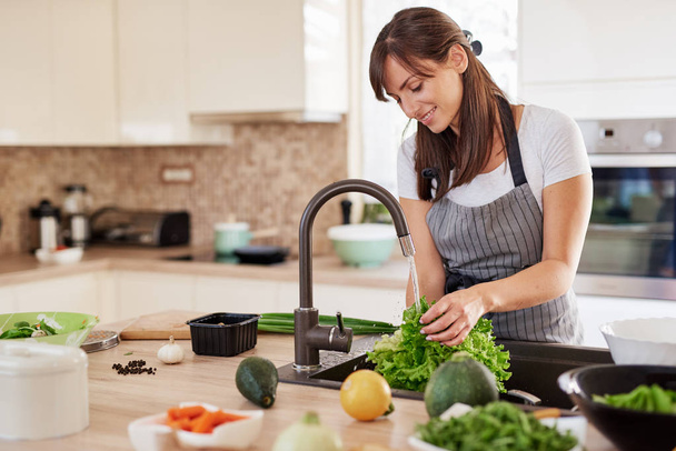 Cheerful attractive Caucasian woman in apron washing salad in kitchen. Dinner at home concept. - Foto, Imagen
