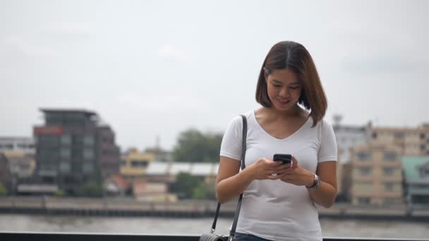 Mulher asiática nova bonita usando o smartphone que gosta de ler as mídias sociais além da cerca no rio
. - Filmagem, Vídeo