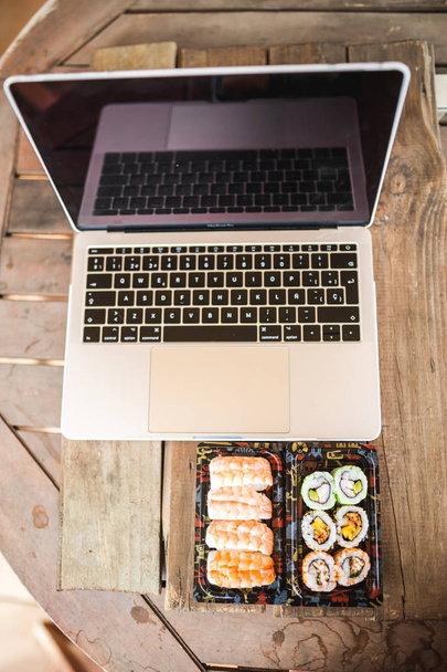 Leckere Sushi-Rollen, Garnelen und Laptop am Holztisch. Essensausgabe. Arbeitsplatz zu Hause - Foto, Bild
