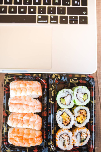 Tasty sushi rolls, shrimp and laptop at wooden table. Food delivery. Workspace at home - Фото, зображення