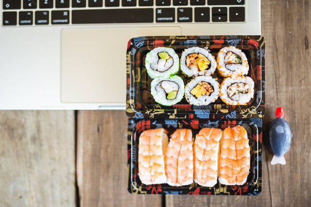 Tasty sushi rolls, shrimp and laptop at wooden table. Food delivery. Workspace at home - Фото, изображение
