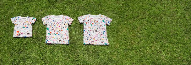 Three T-shirts lying on green grass. Summer camp for children, conceptual. Large family background. Large family background. Advanture summer camp for different aged children. - Photo, Image