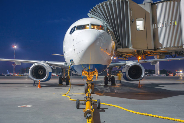 Пасажирський літак припаркований, з'єднаний драбиною в будівлі терміналу в аеропорту, біля передньої причепи лебідки для буксирування трактора вночі
. - Фото, зображення