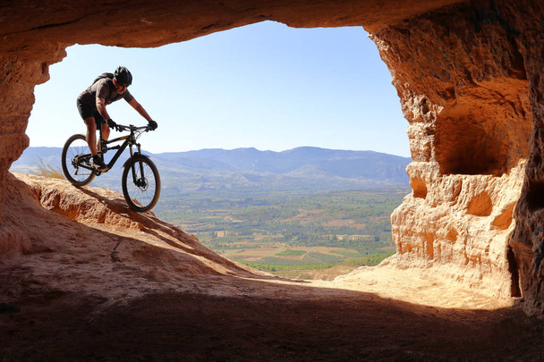 ISOLATED RIDER WITH HELMET AND BACKPACK DESCENDING IN A MOUNTAIN BIKE TOWARDS THE ENTRANCE OF A CAVE IN SUMMER - Photo, Image
