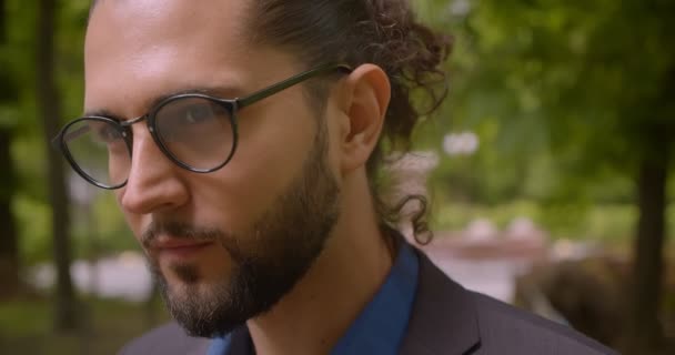 Close-up shot of handsome hipster guy in formal suit with ponytail smiling seductively into camera in park. - 映像、動画
