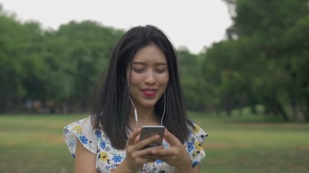 Slow-motion beautiful young Asian woman enjoying listening to music outdoors at a public park. - Footage, Video