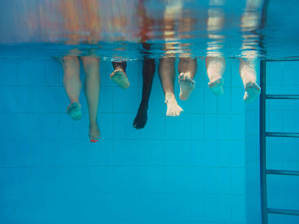 スイミングプールで4人の足が水中に。パーティー。夏。休暇、多様性、友情、スポーツの概念. - 写真・画像