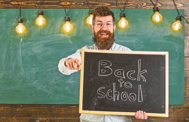 Man with beard and mustache on happy face invites students, pointing forward, chalkboard on background. Teacher in eyeglasses holds blackboard with inscription back to school. Back to school concept - Фото, изображение