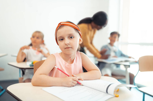 Beautiful cute girl holding pencil while sitting at the desk - 写真・画像