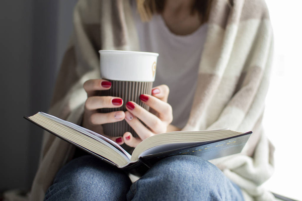 Молодая девушка, покрытая одеялом, сидит дома у окна, читает книгу и пьет чай, копирует место для текста
 - Фото, изображение