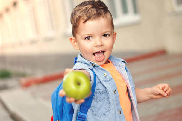 Un niño pequeño de tres años con una mochila en la espalda muestra su manzana verde para un aperitivo
. - Foto, imagen