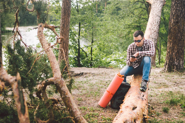 Mladý muž sedí na náklaďáku v lese a používá mobilní telefon. Muž, který sedí na velkém padlým stromě, svíral telefon v ruce. tematikou pro pěší turistiku. Venkovní aktivity jsou pěší. Cestování a dobrodružství - Fotografie, Obrázek