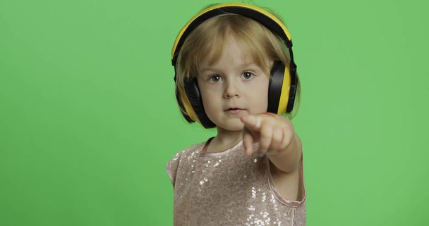 Mädchen in glänzendem Kleid und Kopfhörer hört Musik und zeigt den Finger nach vorne - Foto, Bild