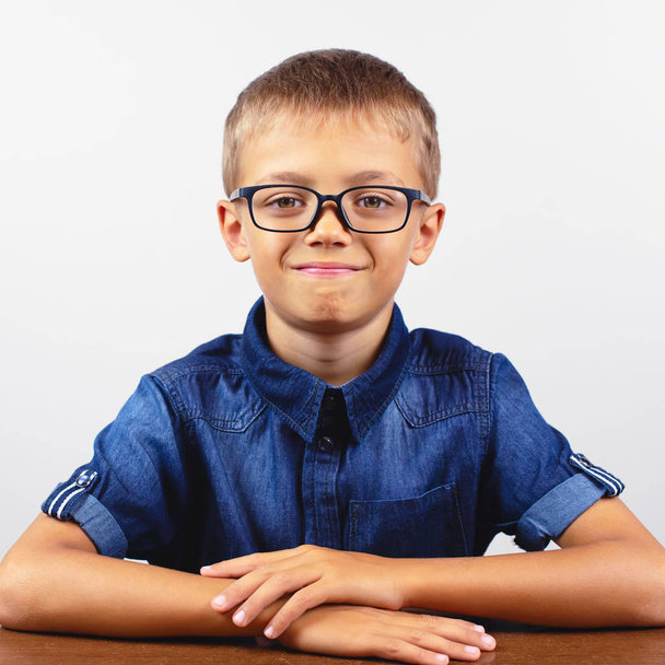 青いシャツを着た男子生徒がテーブルに座っている。白い背景に眼鏡をかけた少年 のコンセプト - 写真・画像