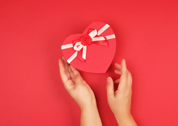 twee handen houden een papier gesloten rode doos in de vorm van een hart  - Foto, afbeelding