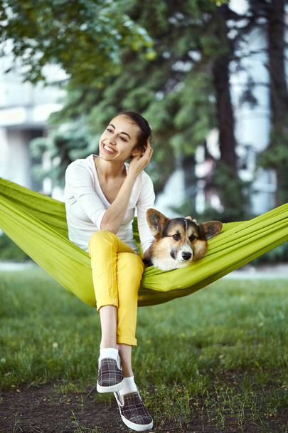 Mujer joven sonriente sentada en hamaca verde con lindo perro galés Corgi en un parque al aire libre. Hermosa hembra feliz en camisa blanca disfrutando de un buen día y acariciando a su perro. Concéntrate en el perro
. - Foto, Imagen
