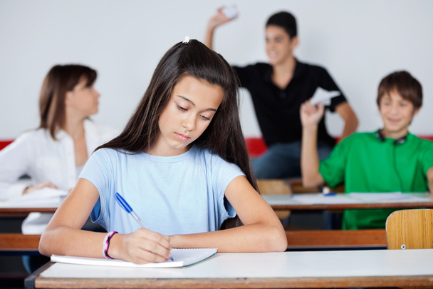 Schoolgirl Writing At Desk While Classmates Playing - Valokuva, kuva