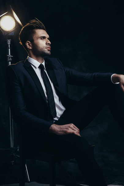 Portret przystojnego mężczyzny siedzącego w studiu z czarnym tłem materialnym i światłem kinowym. - Zdjęcie, obraz
