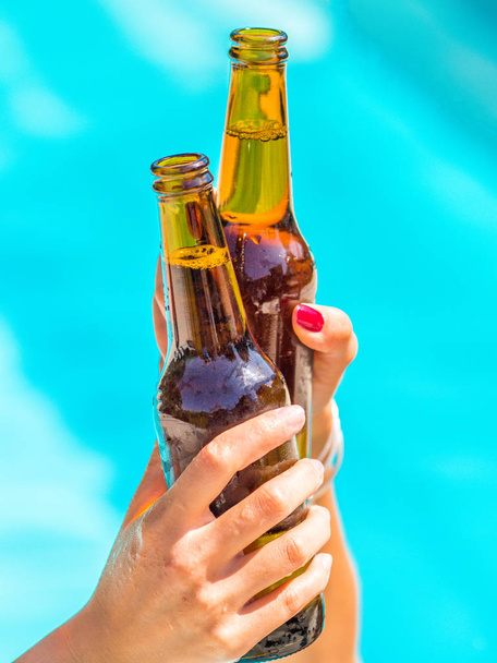 Freundinnen bejubeln klirrende Bierflaschen in den Händen neben einem Pool mit blauem Wasser. - Foto, Bild
