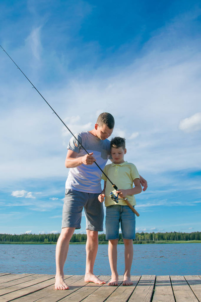 father shows his son on the pier how to keep spinning to catch fish, against a beautiful landscape - Valokuva, kuva