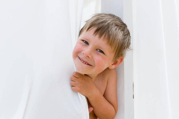 A little boy peeks out from behind a white curtain and smiles. Place for text. - Fotó, kép