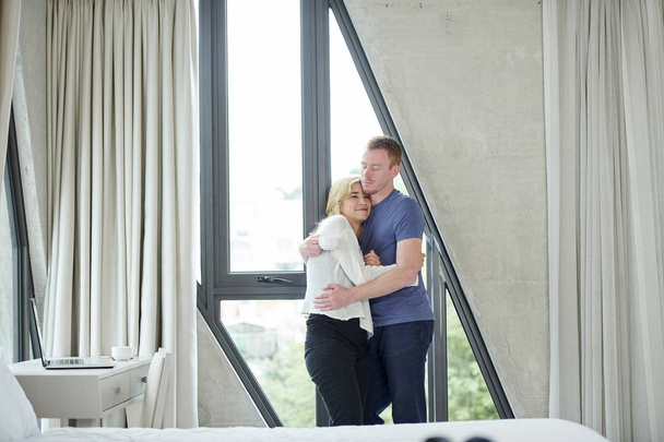 Positivo jovem abraço casal de pé na grande janela em seu apartamento
 - Foto, Imagem