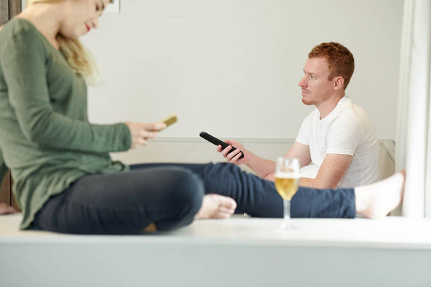 Young man changing channels on tv with remote control when his girlfriend texting friends - Foto, imagen