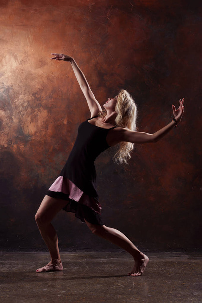 Photo of dancing blonde looking back in black dress on brown background in studio - Foto, afbeelding