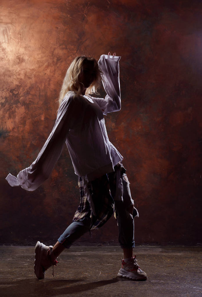 Photo of young dancing blonde looking to side in torn jeans and sneakers on dark brown background - Фото, зображення