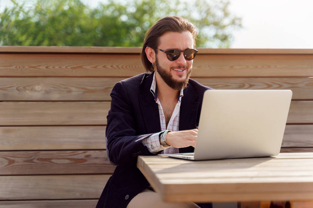Zdjęcie szczęśliwego Brunet człowieka w okularach słonecznych siedzących z laptopem na drewnianej ławce na wiosnę - Zdjęcie, obraz