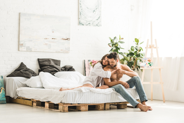 pareja joven abrazándose mientras está sentada en la cama y mirando hacia otro lado
 - Foto, Imagen
