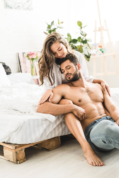 Seksowny człowiek siedzi obok łóżko i dziewczyna przytulanie chłopaka z powrotem - Zdjęcie, obraz