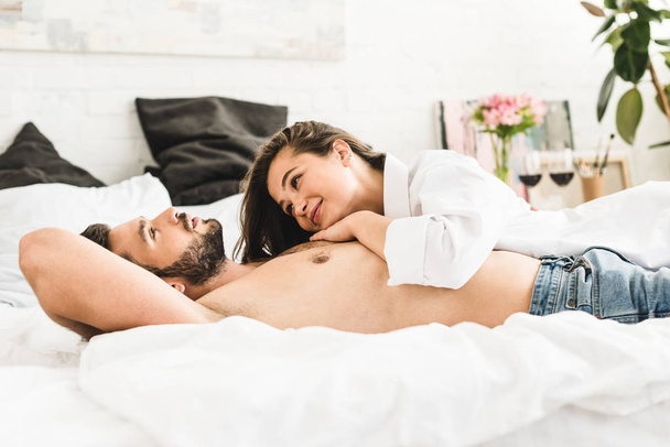Genç çift yatakta yatarken kız gülümseyen ve adam bakıyor - Fotoğraf, Görsel