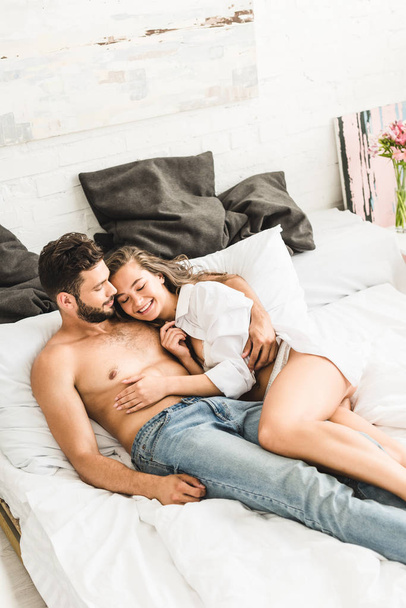 sexy young couple lying in bed and smiling while girl touching shirtless man - Valokuva, kuva