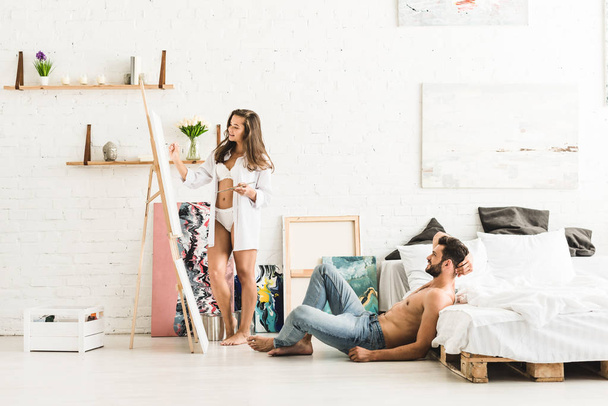 full length view of girl drawing with brush and man sitting on floor near bed - Photo, Image