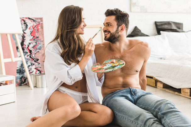 happy couple sitting on floor and looking at each other while girl trying to paint face of guy - Photo, Image